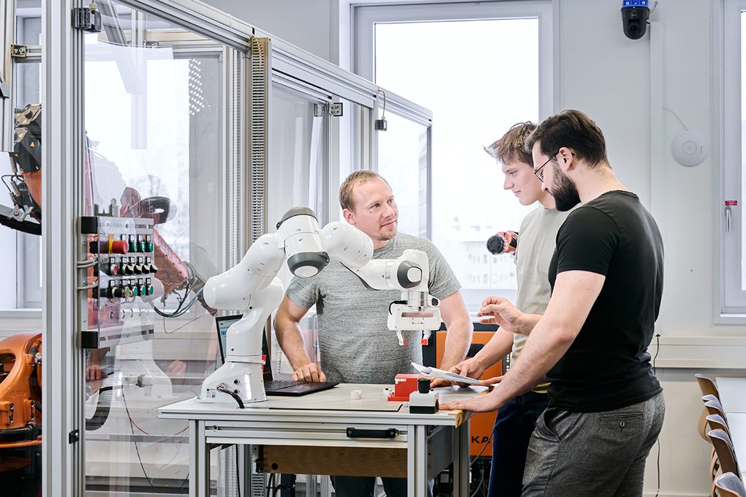Drei Personen besprechen einen Versuchsaufbau mit einem weißen Roboterarm im Labor.