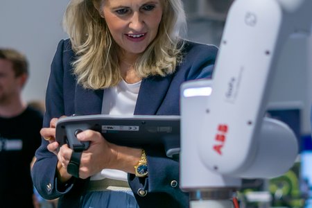 Eine Frau mit blonden Haaren und Tablet schaut auf einen Roboterarm