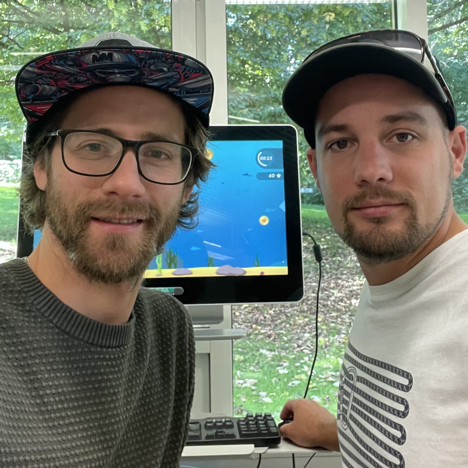 Zwei Männer, beide mit Bärten, der eine mit Brille und Cap, stehen nebeneinander vor einem Bildschirm