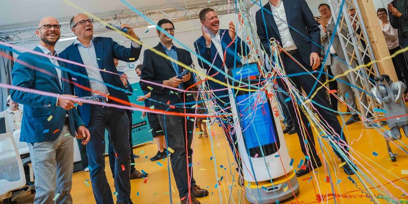 Staatsminister Blume und Forschende stehen vor einem Roboter mit Konfettiregen. 