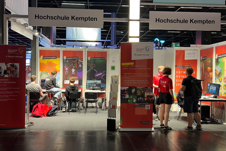 Der Messestand der Fakultät Informatik auf der gamescom mit zwei Rollups im Vordergrund, Bannern an den Messeswänden, davor Tische mit Bildschirmen, an denen Studierende und Messebesucher sitzen.