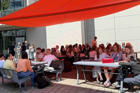 Unter einem orangeroten Sonnensegel sitzen Menschen an mehreren Tischen und unterhalten sich. Links im Hintergrund stehen ein paar Personen. Rechts im Vordergrund hockt eine Person. 