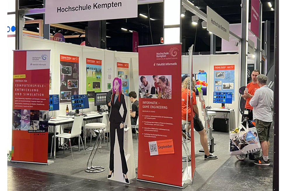 Messestand der Fakultät Informatik mit mehreren Computerplätzen, Postern von Spielen, Werbe-Aufstellern für das Studium und das Zentrum für Computerspiele-Entwicklung. Im Messestand sind Studierende in Fakultäts-T-Shirts und Messebesucher zu sehen.