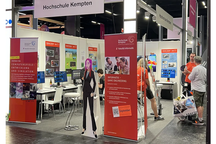 Messestand der Fakultät Informatik mit mehreren Computerplätzen, Postern von Spielen, Werbe-Aufstellern für das Studium und das Zentrum für Computerspiele-Entwicklung. Im Messestand sind Studierende in Fakultäts-T-Shirts und Messebesucher zu sehen.