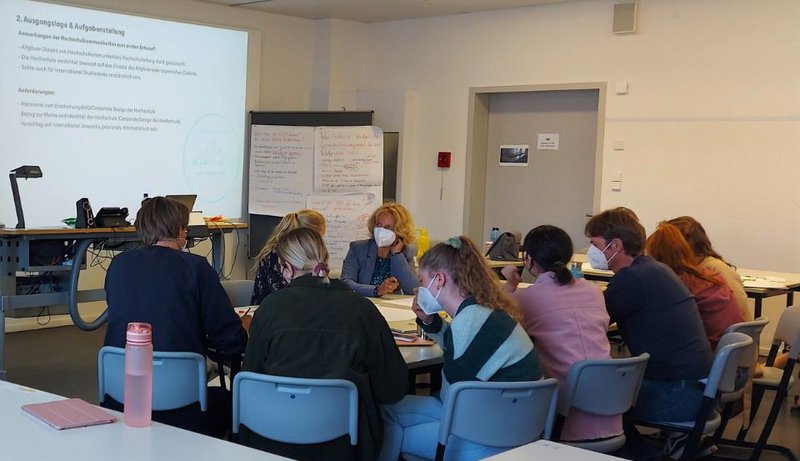 Mehrere Personen sitzen in einem Raum an einem Tisch, größtenteils mit dem Rücken zur Kamera. Sie tragen FFP2-Masken.