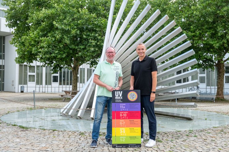 Hochschulpräsident Prof. Dr. Wolfgang Hauke (links) und Thomas Michel, Direktor der AOK Kempten Oberallgäu Lindau (rechts), stehen auf dem Campus der Hochschule Kempten vor einer UV-Index-Tafel. Die Tafel informiert über die aktuelle UV-Belastung und gibt Empfehlungen zum Sonnenschutz. Sie wurde im Rahmen eines gemeinsamen Informationsabends zu den gesundheitlichen Folgen des Klimawandels übergeben und ist Teil des bayernweiten Präventionsprojekts „WATCH OUT“. Im Hintergrund sind die markante Campus-Skulptur sowie Bäume und Hochschulgebäude zu sehen.