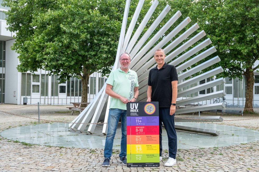 Hochschulpräsident Prof. Dr. Wolfgang Hauke (links) und Thomas Michel, Direktor der AOK Kempten Oberallgäu Lindau (rechts), stehen auf dem Campus der Hochschule Kempten vor einer UV-Index-Tafel. Die Tafel informiert über die aktuelle UV-Belastung und gibt Empfehlungen zum Sonnenschutz. Sie wurde im Rahmen eines gemeinsamen Informationsabends zu den gesundheitlichen Folgen des Klimawandels übergeben und ist Teil des bayernweiten Präventionsprojekts „WATCH OUT“. Im Hintergrund sind die markante Campus-Skulptur sowie Bäume und Hochschulgebäude zu sehen.