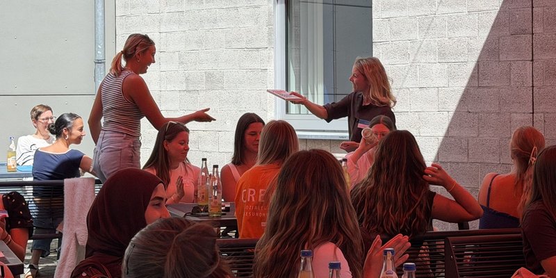 Professorin Carolin Köhler überreicht einer Teilnehmerin des Campustages der Fakultät Soziales und Gesundheit im Rahmen des Ausklangs draußen unter dem roten Sonnensegel ein Buch. 