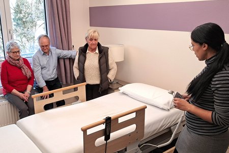 Drei ältere Personen hören einer Frau beim Reden zu. Zwischen ihnen ist ein Aufstehbett zu sehen.