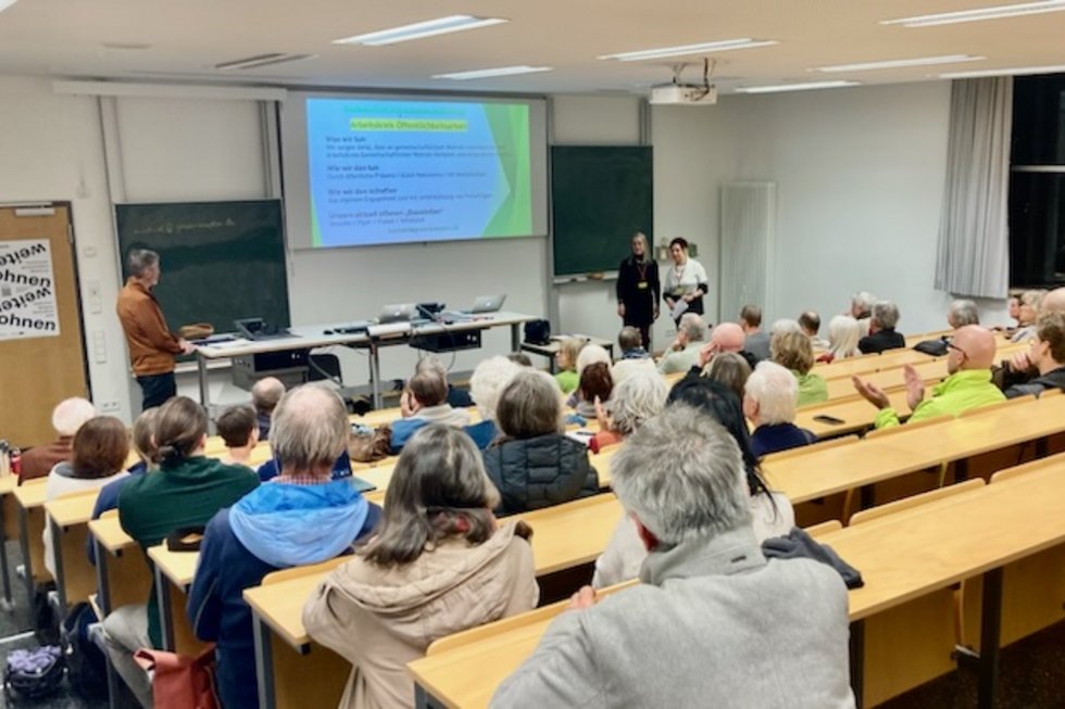 Auf dem Bild ist ein Hörsaal zu sehen mit Menschen aller Altersstufen. Drei Personen halten einen Vortrag mit einer Präsentation, die an die Wand projiziert wird.