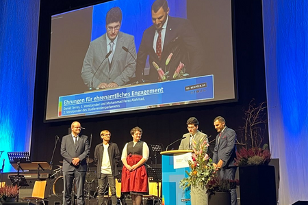 Redner auf der Bühne zeichnet drei Studierende, die daneben stehen, für ihr Ehrenamt an der Hochschule Kempten aus.