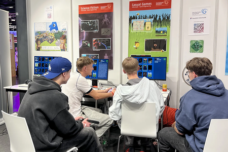 Vier junge Männer sitzen am Messestand vor einem Bildschirm und sehen sich die Spielauswahl der Fakultät Informatik an.