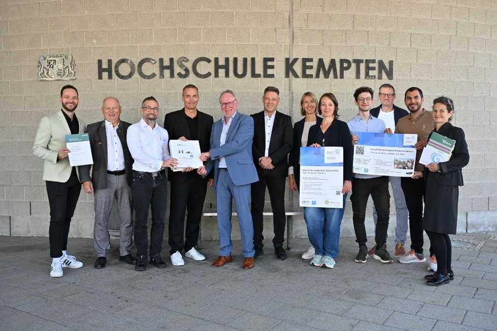 Elf Vertreterinnen und Vertreter der Hochschule Kempten, der AOK Bayern, der Techniker Krankenkasse und der Studierendenvertretung stehen vor dem Schriftzug „Hochschule Kempten“ für ein Gruppenfoto. Hochschulpräsident Prof. Dr. Wolfgang Hauke und Kanzler Christian Herrmann stehen gemeinsam mit den Förderpartnern und dem Team des Hochschulgesundheitsmanagements in der Mitte. Mehrere Personen halten Förderunterlagen und Informationsplakate in den Händen. Anlass ist der Start des neuen Hochschulgesundheitsmanagements, das von AOK Bayern und Techniker Krankenkasse mit insgesamt 300.000 Euro gefördert wird.