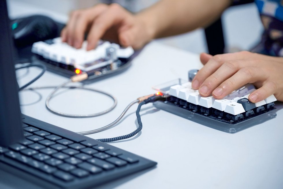 Nahaufnahme zweier Hände, die an einem Computer mit kompakten, programmierbaren Eingabegeräten arbeiten. Die Tastenfelder sind über Kabel mit dem Rechner verbunden und werden parallel bedient. Das Bild steht beispielhaft für die Entwicklung und Erprobung digitaler Anwendungen, benutzerfreundlicher Mensch-Maschine-Schnittstellen sowie innovativer Technologien in Forschung und Lehre an der Hochschule Kempten.