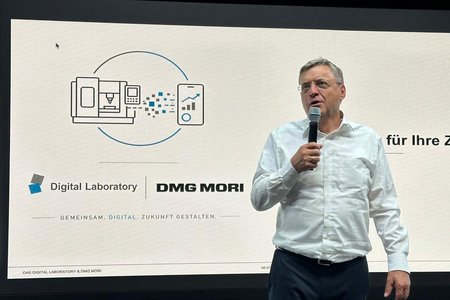 Redner mit Mikrofon vor dem DMG MORI-Logo und einer Leinwand, auf der die Projektpartner stehen: Digital Laboratory und DMG MORI