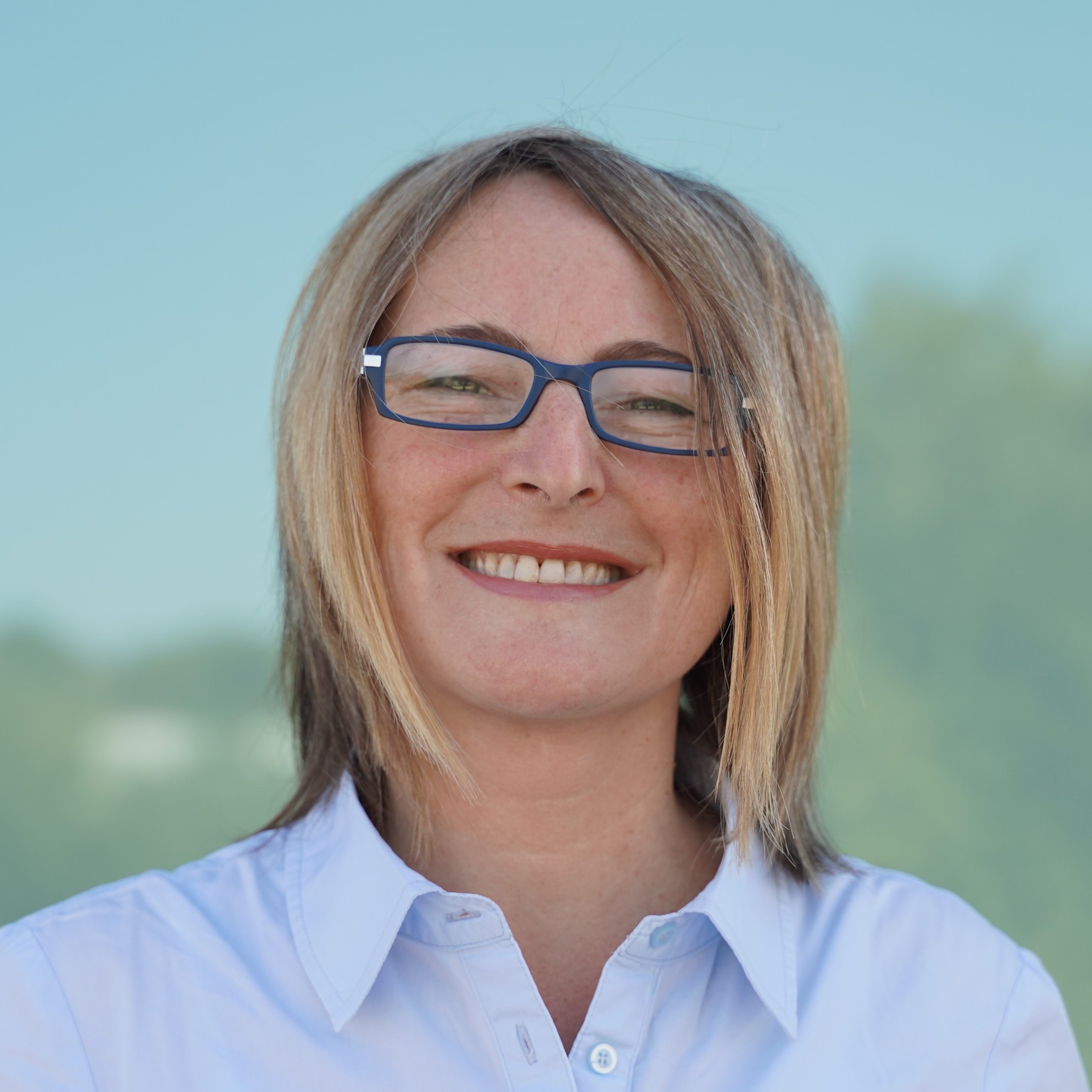 Eine Frau mit kurzen hellbraunen Haaren und blauer Brille lächelt in die Kamera