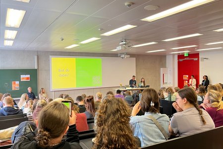 Blick von hinten auf einen gut gefüllten Saal, in dem eine Person spricht. Auf der Leinwand ist eine Präsentation zu sehen. 