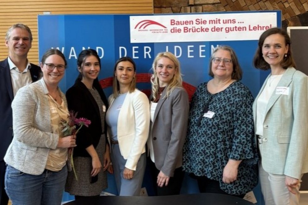 Gruppenfoto von sieben Teilnehmenden am Stand der Hochschule Kempten beim „Bayerischen Tag für Gute Lehre“: Die Personen stehen vor einer blauen Präsentationswand mit der Aufschrift „Wand der Ideen“ und einem Banner zur Lehrinitiative. Im Vordergrund ist ein runder Infotresen mit Logo der Hochschule Kempten zu sehen. Eine Person hält Blumen in der Hand, alle blicken in die Kamera und wirken freundlich und engagiert.