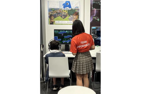 Eine Studierende zeigt einem jungen Messebesucher ein Spiel am Computerbildschirm. Er sitzt mit Headset vor dem Bildschirm.