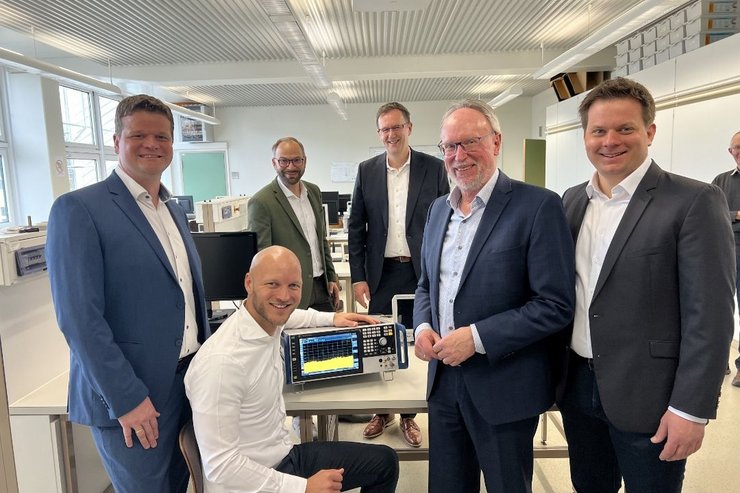 In einem hellen Hochschullabor stehen fünf Männer in Anzügen und ein Mann sitzt vor einem Tisch, auf dem ein eingeschalteter Signal- und Spektrumanalysator von Rohde & Schwarz steht. Der sitzende Mann präsentiert das Gerät und zeigt auf den Bildschirm mit Messkurven. Die Gruppe blickt in die Kamera und wirkt freundlich. Im Hintergrund sind Arbeitsplätze und technische Ausstattung des Labors zu sehen.
