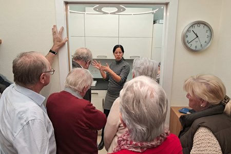 Im Hintergrund erklärt eine Frau etwas. Im Vordergrund sind die Rücken und Köpfe der älteren Zuhörenden zu sehen. 