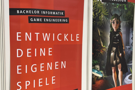 Zwei Banner an den Wänden des Messestands: eines im roten Fakultätsdesign mit dem Schriftzug "Entwickle deine eigenen Spiele", das zweite mit dem Hashtag "Gamedesign" und einem Spielecharakter, einer Art Elfe, im Wald