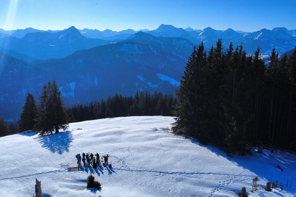 Es ist ein Bergpanorama bei sonnigem Wetter zu sehen. Im Vordergrund ist eine kleinere Gruppe Wanderer im Schnee zu erkennen, die aus der Ferne in die Kamera winken. 