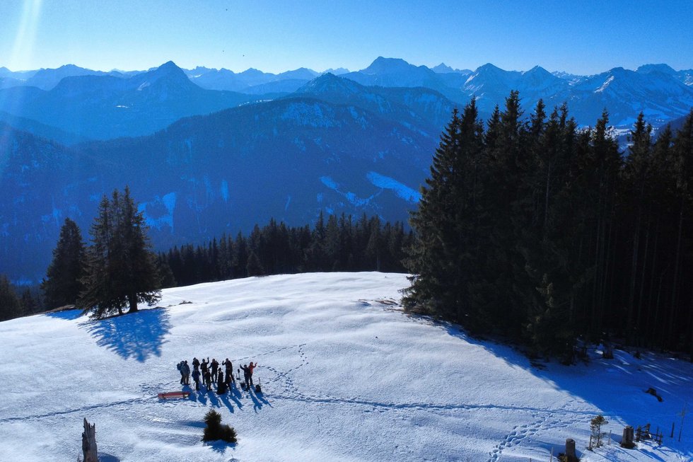 Es ist ein Bergpanorama bei sonnigem Wetter zu sehen. Im Vordergrund ist eine kleinere Gruppe Wanderer im Schnee zu erkennen, die aus der Ferne in die Kamera winken. 