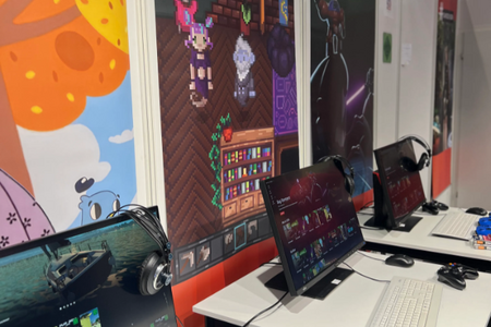 Ausschnitt des Messestands: Drei Computerarbeitsplätze mit Bildschirmen, Controllern, Headsets vor den Bannern, auf denen Szenen unterschiedlicher Spiele zu sehen sind. Auf den Bildschirmen sind die Spiele zu sehen, die die Besucher testen können. 