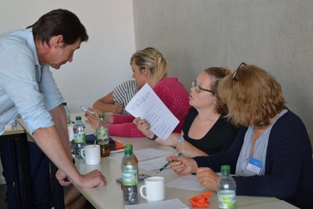 An einem Tisch sitzen mehrere Frauen, ein Mann steht und beut sich zu ihnen herunter. Sie unterhalten sich. 