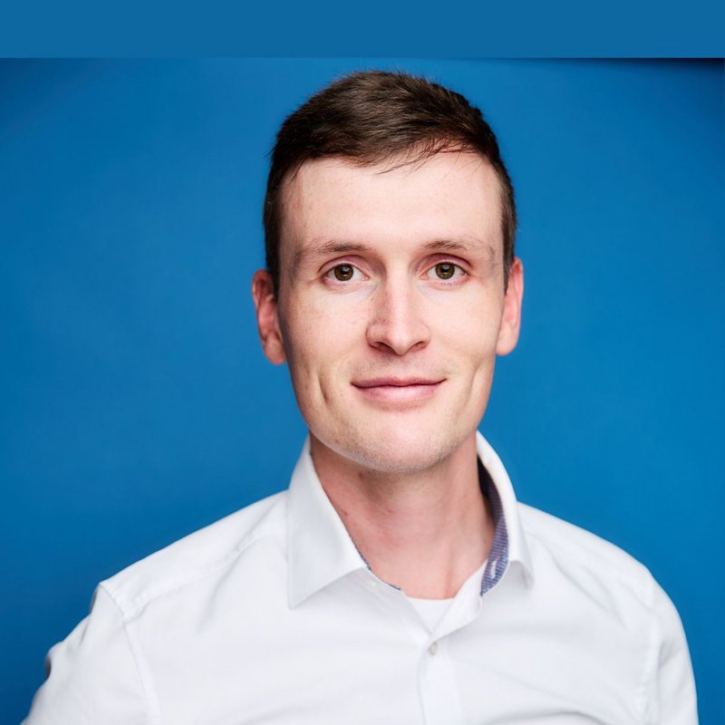 Ein Mann mit braunen Haaren und braunen Augen lächelt in einem weissen Hemd vor einem blauen Hintergrund in die Kamera