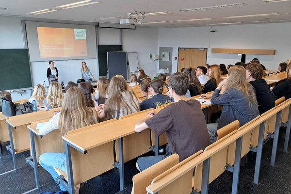 In einem aufsteigenden Hörsaal stehen zwei Dozentinnen und sprechen, während eine Gruppe Jugendlicher zuhört