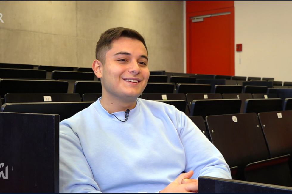 Junger Mann in blauem Pullover sitz in einem Hörsaal mit schwarzen Sitzplätzen