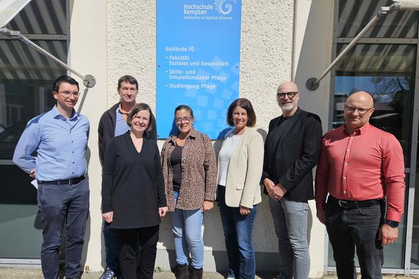 Sieben Frauen und Männer stehen im Sonnenlicht vor einem blauen Schild und schauen in die Kamera.