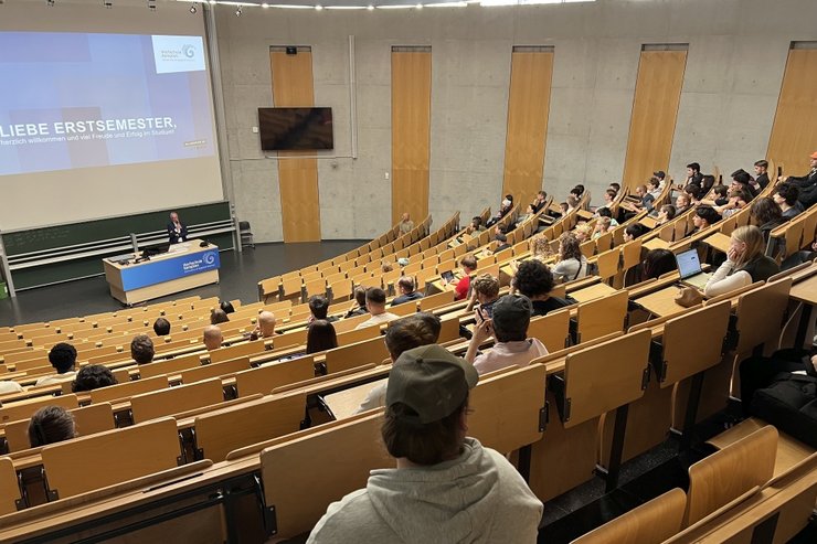 Reihen von Erstsemestern im größten Hörsaal der Hochschule, am Rednerpult der Hochschulpräsident mit dem Begrüßungsscreen auf der Leinwand mit dem Text "Liebe Erstsemster"