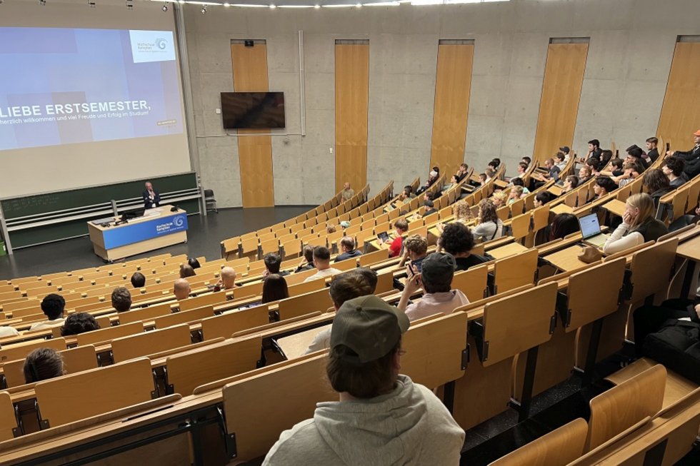 Reihen von Erstsemestern im größten Hörsaal der Hochschule, am Rednerpult der Hochschulpräsident mit dem Begrüßungsscreen auf der Leinwand mit dem Text "Liebe Erstsemster"