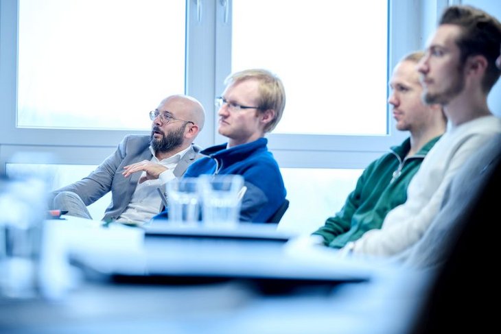 Mehrere Personen sitzen an Besprechungstischen und blicken auf einen Monitor