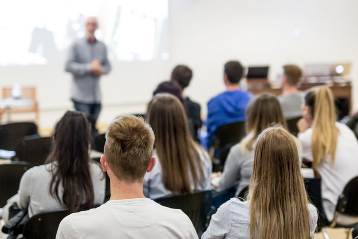 Junge Menschen sitzen in einem Hörsall. Im Vordergrund steht ein Professor vor einer Leinwand.