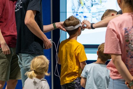 Mehrere Kinder und Erwachsene nehmen beim Tag der offenen Tür des IPI – Institut für Produktion und Informatik an einer Virtual-Reality-Demonstration teil. Ein Mitarbeitender setzt einem Kind eine VR-Brille auf und erklärt die Anwendung, während weitere Kinder gespannt zusehen. Auf einer großen Bildschirmanzeige im Hintergrund wird die virtuelle Umgebung dargestellt. Die Szene zeigt, wie das Institut Forschung und digitale Technologien für Besucherinnen und Besucher erlebbar macht.