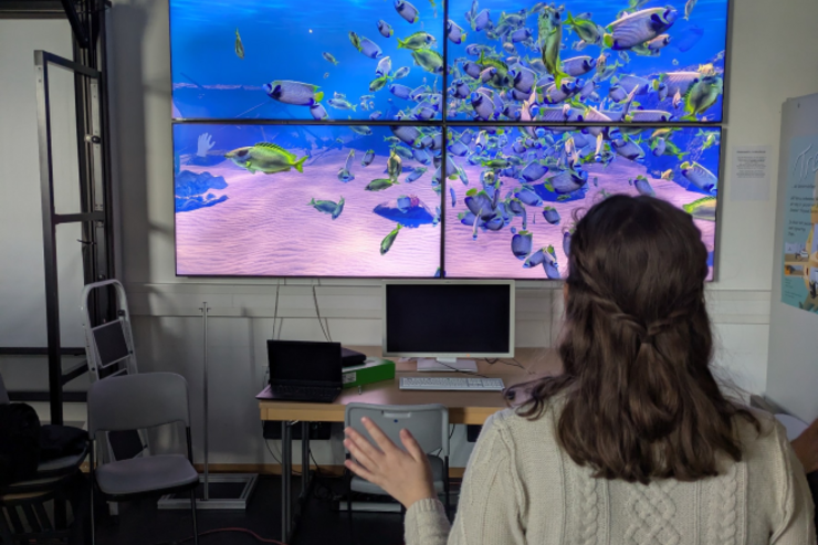 Bildschirmwand im GamesLab, bestehend aus vier Screens mit einer simulierten Unterwasserwelt. Die Tiere darin werden durch Gesten einer Frau gesteuert, die vor der Bildschirmwand steht und eine Hand hebt.