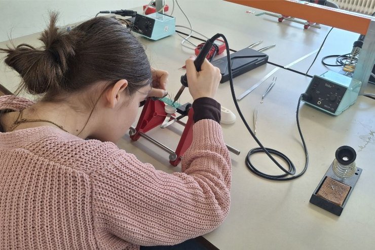 In einem Techniklabor arbeitet eine Schülerin konzentriert an einer elektronischen Aufgabe und lötet mit einem Lötkolben ein Bauteil in einer Haltevorrichtung. Auf dem Tisch liegen weitere Werkzeuge und Geräte wie Lötstation, Kabel und Messinstrumente. Die Szene vermittelt praktisches Arbeiten und Einblicke in technische Ausbildung oder ein Mitmachangebot wie den Girls’ Day.