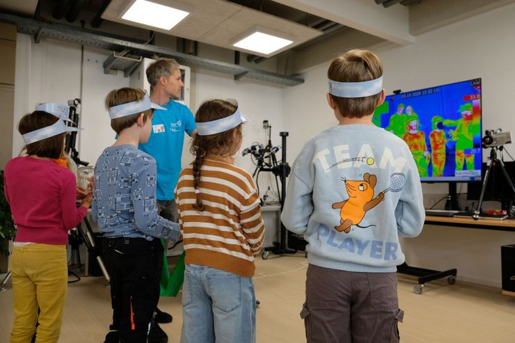 Mehrere Kinder stehen bei „Türen auf mit der Maus“ in einem Labor der Fakultät Maschinenbau der Hochschule Kempten und beobachten eine Wärmebildaufnahme auf einem großen Bildschirm. Ein Mitarbeiter erklärt die Technik und die unterschiedlichen Temperaturbereiche, die farbig dargestellt werden. Die Kinder tragen Papierstirnbänder mit Maus-Motiv und erleben so spielerisch, wie technische Kameras Wärme sichtbar machen.