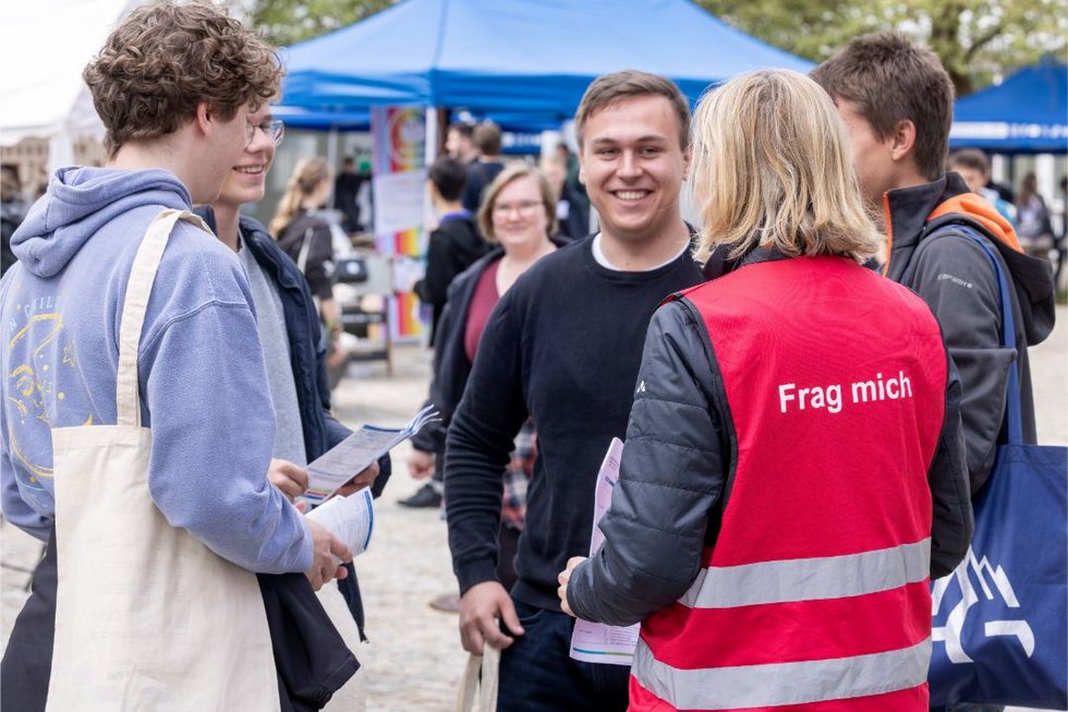 Auf dem Gelände der Allgäuer Hochschulmesse stehen mehrere Studierende oder Studieninteressierte im Gespräch mit einer Person, die eine rote Weste mit der Aufschrift „Frag mich“ trägt. Die Gruppe wirkt offen und interessiert, einige halten Flyer oder Broschüren. Im Hintergrund sind Stände und Pavillons der Veranstaltung zu sehen, die eine lebendige Messeatmosphäre vermitteln.