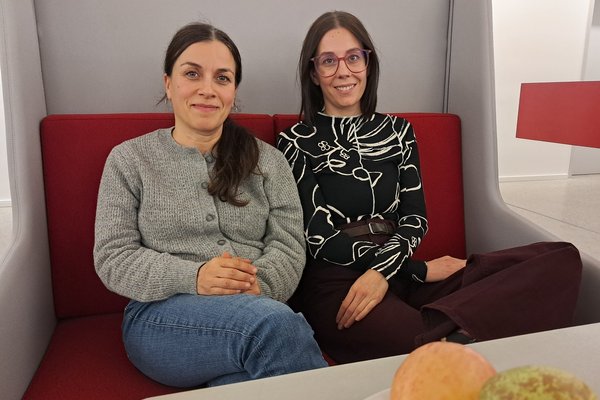 Prof. Dr. Gökçen Yüksel und Prof. Dr. Roxana Bettingersitzen in einem Innenraum auf einer Bank aus Beton mit rotem Polster und schauen in die Kamera.