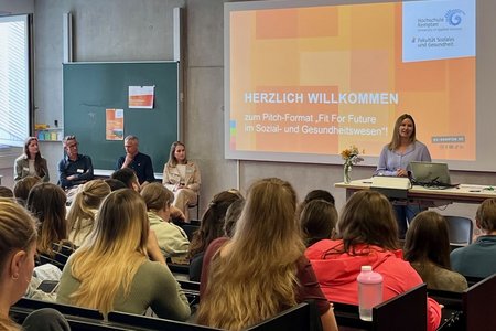 Blick von hinten auf einen gut gefüllten Saal, in dem eine Person spricht. Auf der Leinwand ist eine Präsentation zu sehen. 
