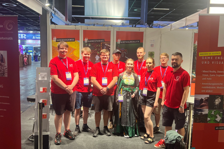 Gruppenfoto des Standpersonal im Messestand. Zu sehen sind Prof. Dr. Bühling und acht Studierenden, davon eine im Cosplay-Kostüm, die anderen in den roten Fakultäts-T-Shirts.