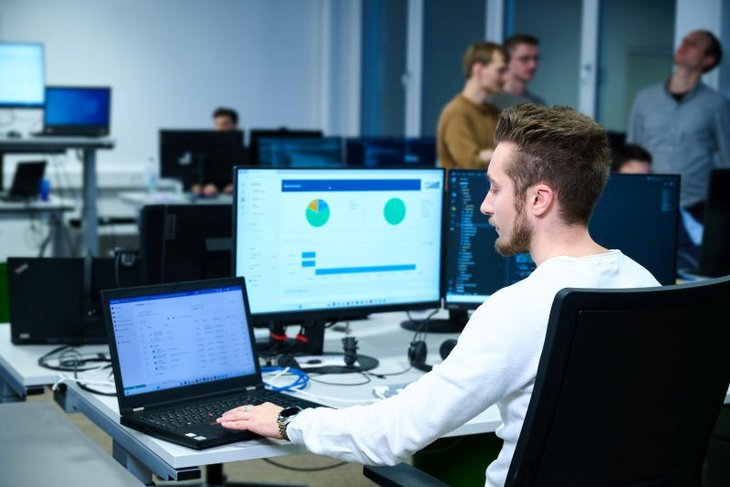 Mitarbeiter sitzt am Schreibtisch im Großraumbüro mit einem Laptop und einem Monitor, auf dem schemenhaft Tortendiagramme zu sehen sind.