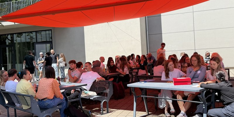 Die Teilnehmenden des Campustages sitzen vor der Mensa draußen gemütlich bei schönen Wetter unter unserem roten Sonnensegel gemütlich beisammen.